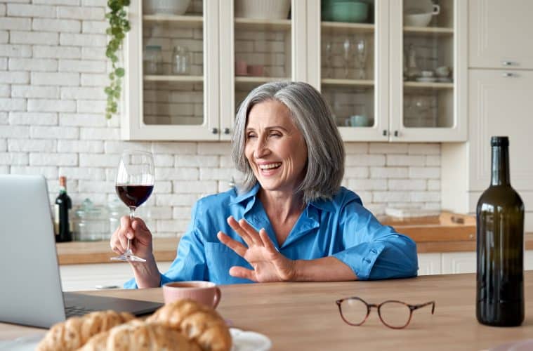 Happy 60s mature woman drinking wine video calling friend on laptop at home.